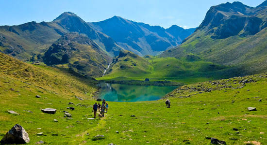 Walkers crossing a green Pyrenean valley beside a mountain lake